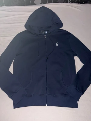 Mörkblå hoodie från Polo Ralph Lauren - Snygg mörkblå hoodie från Polo Ralph Lauren med dragkedja, två fickor fram och klassisk vit broderad logga på bröstet. Tillverkad i mjukt bomullsmaterial och har justerbar huva med snören. Perfekt för en chill och stilren look.