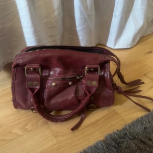 Vinröd handväska med nitar - Snygg vinröd handväska i skinnimitation med detaljer i mässingsfärgad metall och dekorativa nitar. Köpt i Italien för 500kr och säljer för 350, kan självklart gå ner i pris vid snabb affär❤️Band medkommer!