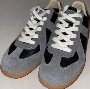 Maison Margiela svarta och grå sneakers - Snygga sneakers från Maison Margiela i svart läder med grå mockadetaljer och beige/brun sula. Klassisk låg modell med vita snören och stilren design som passar till det mesta. Perfekta för dig som gillar exklusiva och trendiga skor.