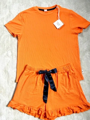 Chelsea Peers orange pyjamasset - Superfint orange pyjamasset från Chelsea Peers i ribbad mjuk bomull. Setet består av en kortärmad t-shirt och shorts med volangkant och svart sidenband i midjan. Perfekt för mysiga kvällar hemma.