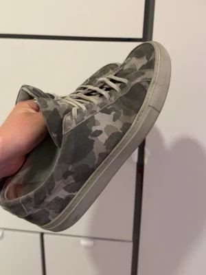 Common projects camosneakers - Snygga sneakers i grått camomönster med ovandel i mocka och vita snören. Skorna har en klassisk låg siluett och vit sula som ger en clean look. Perfekta för dig som vill ha en streetstyle-vibe i vardagen. Nypris 4000kr mitt pris 799
