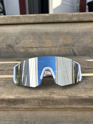 Vita sportiga spegelglas-solglasögon - Säljer ett par sportiga solglasögon med vit båge och stora spegelglas i silver. Glasögonen har en futuristisk, kantig design och bred lins som täcker mycket av ansiktet. Perfekta för dig som gillar en modern och cool stil. En liten repa som ej syns vid användning 
