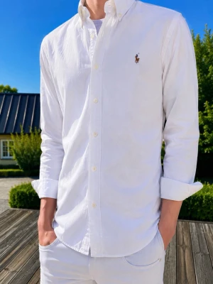 Vit skjorta från Ralph Lauren - (MODDELEN ÄR 185 och väger 75kg)Klassisk vit skjorta från Ralph Lauren med button-down krage och broderad logga på bröstet. Skjortan har lång ärm, normal passform och är tillverkad i mjuk bomull. Perfekt för en clean och stilren look.