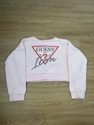 Ljusrosa cropped hoodie från Guess - Säljer en ljusrosa cropped hoodie från Guess med stort tryck framtill i rött, svart och vitt. Ikonisk trekantslogga och texten 'Icon' i glittrigt mönster. Tröjan har långa ärmar, ribbade muddar och är gjord i mjukt bomullsmaterial. Perfekt för en trendig och avslappnad look.