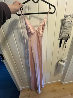 Ljusrosa långklänning/balkläning - Säljer en elegant ljusrosa långklänning/balkläning i satin med smala axelband och v-ringning. Klänningen har öppen rygg med en fin knytning och en mjukt följsam passform som ger en snygg siluett. Perfekt för dig som vill ha en stilren och trendig look. Den är från SHEIN och skulle ha den på min bal men blev en annan tyvärr. Har bara provat den.