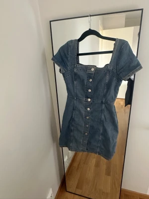 Kort jeansklänning med knappar - Snygg kort jeansklänning med markerad midja och knappar längs hela framsidan. Klänningen har korta ärmar och fyrkantig urringning. Perfekt för dig som gillar en avslappnad men trendig stil.