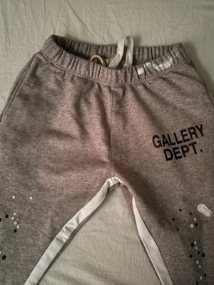 Grå mjukisbyxor från Gallery Dept. - Grå mjukisbyxor från Gallery Dept. med svart logga på ena benet, vita snören i midjan och vita detaljer längs benen. Byxorna har elastisk midja och är tillverkade i 100% bomull. Snygga färgstänk och slitningar för en trendig look.