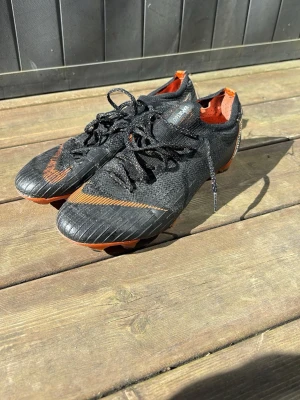 Nike Mercurial Vapor 360 fotbollsskor - Säljer ett par Nike Mercurial Vapor ACC fotbollsskor i svart med orange detaljer och logga. Skorna har låg profil, snörning och ovandel i textil/syntet för bra passform och känsla på planen. Perfekta för dig som vill ha snabba och lätta skor till match eller träning. Äldre modell som är väldigt sällsynt men har en liten defekt som visat på bild. Går att spela normalt känns ej av på något sätt vara utseendet som lurar.