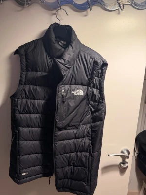 Svart dunväst från The North Face - Hej säljer en Snygg svart dunväst från The North Face med hög krage och dragkedja. Västen har en quiltad design, logga på bröstet och ryggen samt en praktisk bröstficka med dragkedja. Perfekt för lager-på-lager och håller dig varm under kyliga dagar.