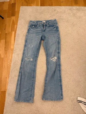 Ljusblå bootcut jeans med slitningar - Säljer ett par ljusblå bootcut jeans med slitna detaljer och hål på benen. Jeansen har klassisk femficksmodell, låg midja och råa kanter vid bensluten. Perfekta för en avslappnad och trendig look.