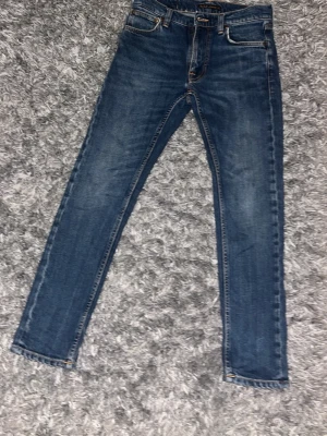Blå slim fit jeans från Nudie Jeans - Säljer ett par klassiska blå slim fit jeans från Nudie Jeans med snygga slitningar och kontrastsömmar. Jeansen har fem fickor, normal midja och är tillverkade i mjuk denim. Perfekta för en avslappnad och trendig look.