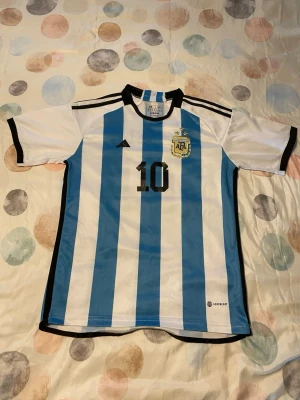 Argentina matchtröja Adidas nr 10 - Säljer en klassisk Argentina fotbollströja från Adidas med blå och vita vertikala ränder, svart nummer 10 på bröstet och AFA-emblemet broderat. Tröjan har svarta detaljer vid krage och ärmslut samt tre svarta ränder på axlarna. Tillverkad i lätt och ventilerande material. STORLEK BARN JUNIOR 15-16 ÅR!!!  170CM