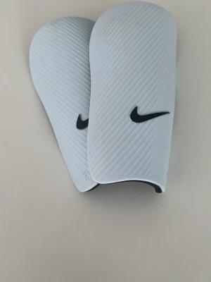 Vita benskydd från Nike - Säljer ett par stilrena vita benskydd från Nike med svart logga. De har en lätt böjd form för att sitta skönt på benet och är tillverkade i ett tåligt plastmaterial med räfflad yta. Perfekta för fotbollsträning eller match.