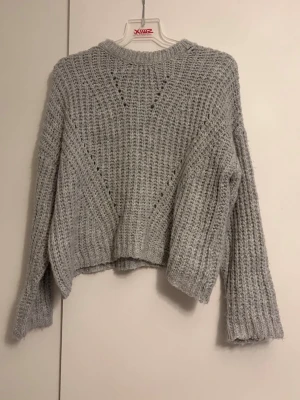Ljusgrå stickad tröja med struktur - Mysig ljusgrå stickad tröja med avslappnad passform och ribbade muddar. Tröjan har ett snyggt strukturmönster med håldetaljer framtill och rund halsringning. Perfekt för lager på lager och chill dagar. Materialet känns mjukt och fluffigt.