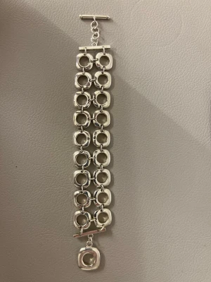 Silverfärgat armband med ringar - Snyggt armband i silverfärg med tre rader av stora, runda länkar och en unik fyrkantig detalj som hänger längst ner. Armbandet har ett T-spänne och en modern, chunky design som verkligen sticker ut. Perfekt för dig som gillar statement-smycken.