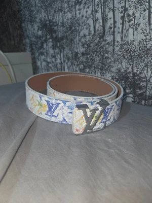 Flerfärgat Louis Vuitton bälte - Louis Vuitton bälte med vit bas och färgglatt LV-mönster i blått, gult och orange. Bältet har ett stort, silverfärgat LV-spänne och är gjort i skinn. Perfekt för att lyfta vilken outfit som helst.