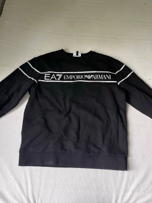 Svart EA7 Emporio Armani sweatshirt - Säljer en svart sweatshirt från EA7 Emporio Armani med vit logga och text framtill. Tröjan har rund hals, långa ärmar och vita detaljer längs axlarna. Perfekt för dig som gillar stilrena och exklusiva plagg.