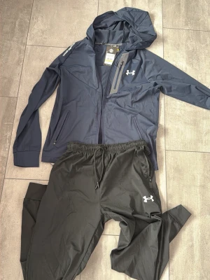Under Armour träningsset marinblå/svart - Snyggt träningsset från Under Armour med marinblå zip-hoodie och svarta byxor. Jackan har huva, dragkedja, bröstficka med dragkedja och logga på bröstet. Byxorna har resår i midjan och logga på benet. Perfekt för löpning eller gym.