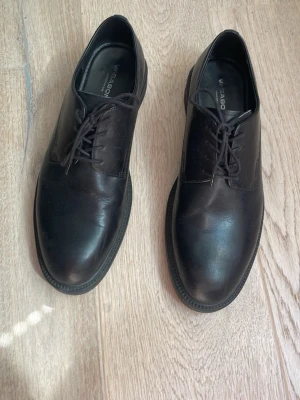 Svarta kostymskor från Vagabond - Stilrena svarta skor från Vagabond i slätt läder. Klassisk design med rundad tå och snörning framtill. Passar perfekt till både jeans och kostym för en clean look. Snygga detaljer och robust sula för extra komfort. Passar 43, använd 1 gång.