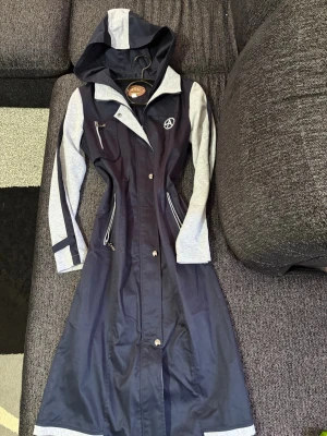 Lång marinblå hoodie-kappa Dooka - Snygg lång marinblå kappa från Dooka med gråa ärmar och detaljer. Kappan har huva, dragkedjor på fickorna och en cool patch på bröstet. Figurnära passform och sportig look med kontrastfärgade inslag. Perfekt för dig som vill sticka ut.