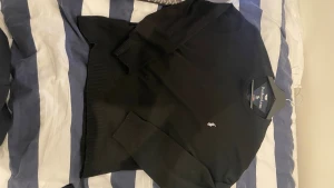 Svart stickad tröja från Polo Ralph Lauren - Klassisk svart stickad tröja från Polo Ralph Lauren med rund halsringning och den ikoniska broderade loggan i vitt på bröstet. Tröjan har långa ärmar och ribbade muddar vid ärmslut och nederkant. Perfekt för en stilren look.