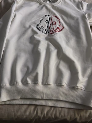 Vit sweatshirt från Moncler - Säljer en stilren vit sweatshirt från Moncler med broderad logga i rött och blått på bröstet. Tröjan har rund halsringning, ribbade muddar och är tillverkad i mjukt material som känns skönt mot huden. Perfekt för dig som gillar exklusiva streetwear-plagg.