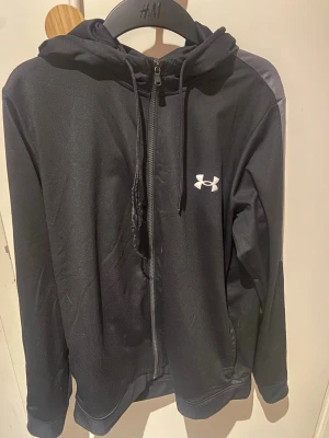 Under armour zip tröja svart - Snygg svart hoodie från Under Armour med dragkedja framtill och justerbar huva med snören. Tillverkad i ett mjukt och sportigt material, perfekt för chill eller träning. Ikonisk logga på bröstet i vitt ger en clean look.
