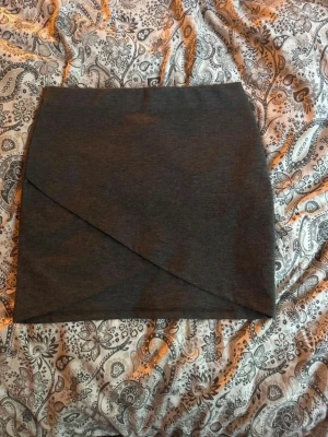 Mörkgrå omlottkjol - Snygg mörkgrå kortkjol med omlottdetalj framtill. Kjolen har en enkel och stilren design som passar perfekt till många olika outfits. Tillverkad i mjukt material för skön passform.