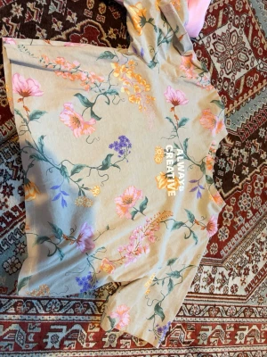 Beige blommig topp med tryck - Trendig beige topp med färgglatt blommönster i rosa, lila och gult. Trycket 'ALWAYS CREATIVE' framtill ger en cool touch. Toppen har korta ärmar och rak passform, perfekt för en avslappnad stil.