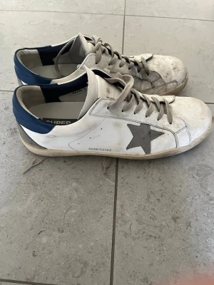 Golden Goose vita sneakers med blå häl - Golden Goose sneakers i vitt läder med grå stjärna på sidan och blå detalj vid hälen. Klassisk låg modell med beige snören och slitna detaljer för en cool, avslappnad look. Snygga till jeans eller shorts och perfekta för dig som gillar streetstyle. Tvättas innan post 