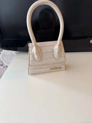 Jacquemus handväska beige i croc-mönster, axelrem ingår - Säljer en superfin liten handväska från Jacquemus i beige färg med croc-mönstrad struktur. Väskan har ett stilrent och trendigt utseende med ett böjt handtag och guldfärgad Jacquemus-logga. AXELREM ingår.