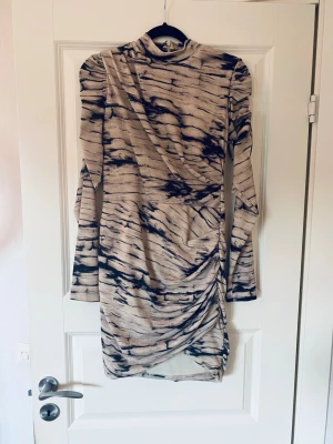 Beige och svart mönstrad kortklänning - Trendig kortklänning i beige med svart abstrakt mönster. Klänningen har lång ärm, hög hals och snygg rynkning på sidan för en cool look. Perfekt för dig som gillar att sticka ut. Mer som en XS i storlek.