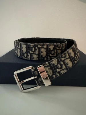 Svart och beige Dior bälte - Snyggt bälte från Dior med ikoniskt monogrammönster i svart och beige. Bältet har ett silverfärgat spänne med Dior-logga och är tillverkat i textil med exklusiv känsla. Perfekt accessoar för att lyfta din outfit.