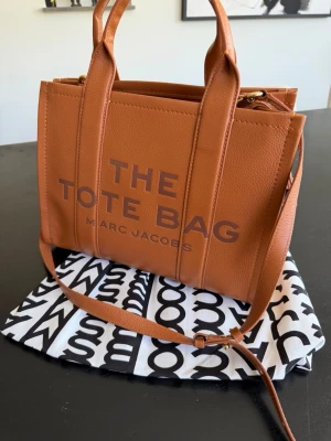 Brun The Tote Bag från Marc Jacobs - Säljer en snygg brun handväska från Marc Jacobs, modellen The Tote Bag. Väskan är i äkta läder med både handtag och justerbar axelrem. Rymligt huvudfack med innerficka och dragkedja. 