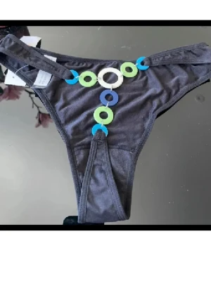 Bikini underdel  med färgglada ringar - Snygga bikini underdel från Lindex med dekorativa ringar i blått, grönt och vitt framtill. Trosorna har låg midja och är gjorda i mjukt stretchigt material för en bekväm passform. Perfekt för dig som gillar unika detaljer och vill ha något extra i