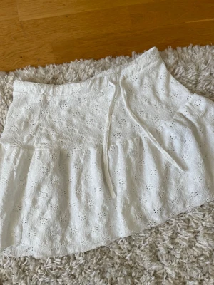 Vit broderad kjol från 157 - Supersöt vit kjol från 157 i storlek XS. Kjolen har broderade blommor, volang och dragsko i midjan för en skön passform. Perfekt till sommaren och ger en fräsch och romantisk vibe till din outfit.