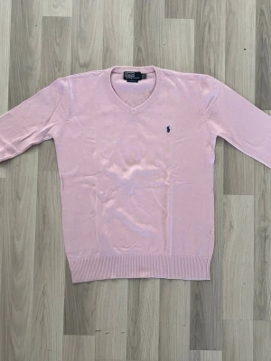 Ljusrosa tröja Polo Ralph Lauren - Ljusrosa tröja från Polo Ralph Lauren med v-ringning och blå broderad logga på bröstet. Tröjan har ribbade muddar vid ärmslut och nederkant och är tillverkad i mjuk bomull för en klassisk och stilren look.