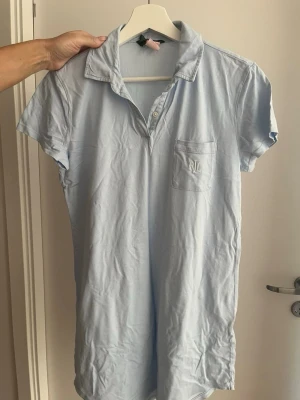 Ljusblå kort pikéklänning från Ralph Lauren - Ljusblå kortärmad pikéklänning från Ralph Lauren med klassisk krage, knappar framtill och bröstficka med RL-broderi. Tillverkad i mjuk bomull, perfekt för en avslappnad och stilren look.