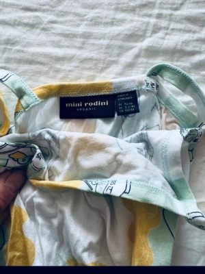 Mini Rodini mönstrad sommarklänning - Söt klänning från Mini Rodini i ekologisk bomull med smala axelband och knyt i midjan. Klänningen har ett lekfullt mönster i ljusgrönt, vitt och gult med tecknade figurer. Perfekt för varma dagar och ger en avslappnad vibe.
