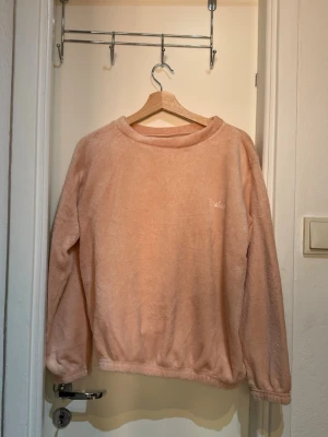 Mjuk rosa loungewear - Supermysigt loungewear-set i ljusrosa fleece. Setet består av en långärmad tröja med rund hals och matchande byxor med resår i midja och benslut. Perfekt för chill hemma eller mysiga kvällar. Broderad logga på bröstet och benet.