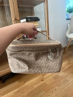 Beige necessär från Guess med logga - Säljer en beige necessär från Guess med klassiskt logomönster och dragkedja. Väskan har ett praktiskt handtag upptill och flera fack inuti för smidig organisering av dina saker. Perfekt för smink eller småprylar när du är på språng.