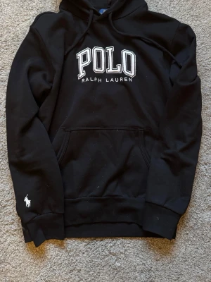 Svart hoodie från Polo Ralph Lauren - Säljer en svart hoodie från Polo Ralph Lauren med stor broderad logga på bröstet och klassisk känguruficka. Tröjan har huva med dragsko och en liten vit broderad häst på ärmen. Perfekt för en chill och stilren look.