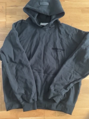 Svart Essentials Fear of God hoodie - Säljer en svart hoodie från Essentials Fear of God med stor logga på ryggen och mindre logga på bröstet. Hoodien har en klassisk känguruficka, ribbade muddar och en rymlig huva. Tillverkad i mjukt bomullsmaterial som är perfekt för en avslappnad streetwear-look.