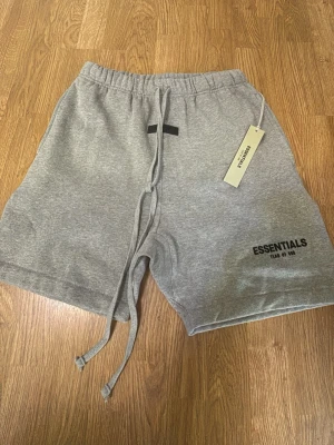 Grå Essentials shorts med snörning - Snygga grå shorts från Essentials med elastisk midja och långa snören framtill. Mjuk bomullskänsla och tryckt logga på benet. Perfekta för chill dagar eller träning. Klassisk loose fit och enkel design.
