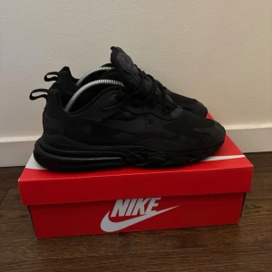 Svarta Nike Air Max 270 React - Snygga helsvarta Nike Air Max 270 React sneakers med chunky sula och diskret logga på plösen. Skorna har en sportig siluett, mesh- och syntetmaterial samt synlig Air-enhet i hälen för extra komfort. Perfekta för dig som gillar stilrena och moderna sneakers.