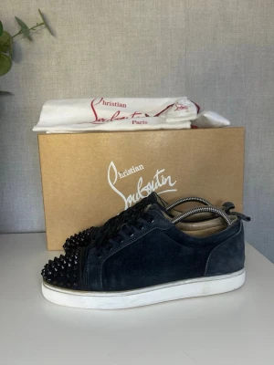Navy Christian Louboutin sneakers - Säljer ett par Navy sneakers från Christian Louboutin i mocka med coola svarta nitar på tån. Skorna har vita sulor och klassisk låg profil. Perfekta för dig som vill sticka ut med en edgy och lyxig stil. Defekter syns på bilderna. Og medföljer endast om vi pratat om det. Size 40 passar 41/42