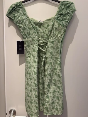 Grön blommig kortklänning med snörning - Söt ljusgrön kortklänning med blommigt mönster och puffärmar. Klänningen har dragkedja i sidan och är tillverkad i ett mjukt bomullsliknande material. Perfekt för sommarens alla tillfällen.