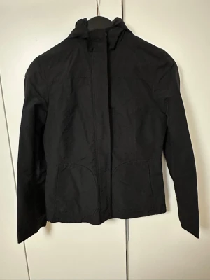 Svart vindjacka från H&M med huva - Snygg svart vindjacka från H&M med huva och dragkedja framtill. Jackan har två stora fickor och en enkel, stilren design. Tillverkad i ett lätt och vindtätt material som passar perfekt för blåsiga dagar.