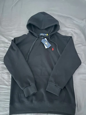 Svart hoodie från Polo Ralph Lauren - Snygg svart hoodie från Polo Ralph Lauren med klassisk huva och dragsko. Tröjan har det ikoniska röda Polo-märket broderat på bröstet och ribbade muddar vid ärmar och nederkant. Tillverkad i mjukt material för en skön och avslappnad stil.