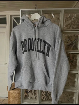 Grå hoodie pull&bear - Snygg grå hoodie med stor svart BROOKLYN-text på bröstet. Klassisk känguruficka framtill och justerbar huva med snören. Mjuk och bekväm, perfekt för en avslappnad streetwear-look.